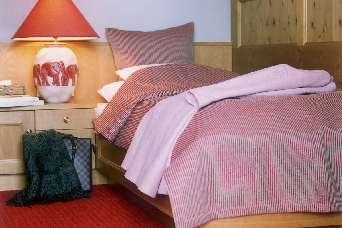 Une chambre d’hôtel avec un lit en bois, une couvre-lit à rayures rouges et blanches, une lampe rouge et une couverture à motifs verts posée sur le sol.