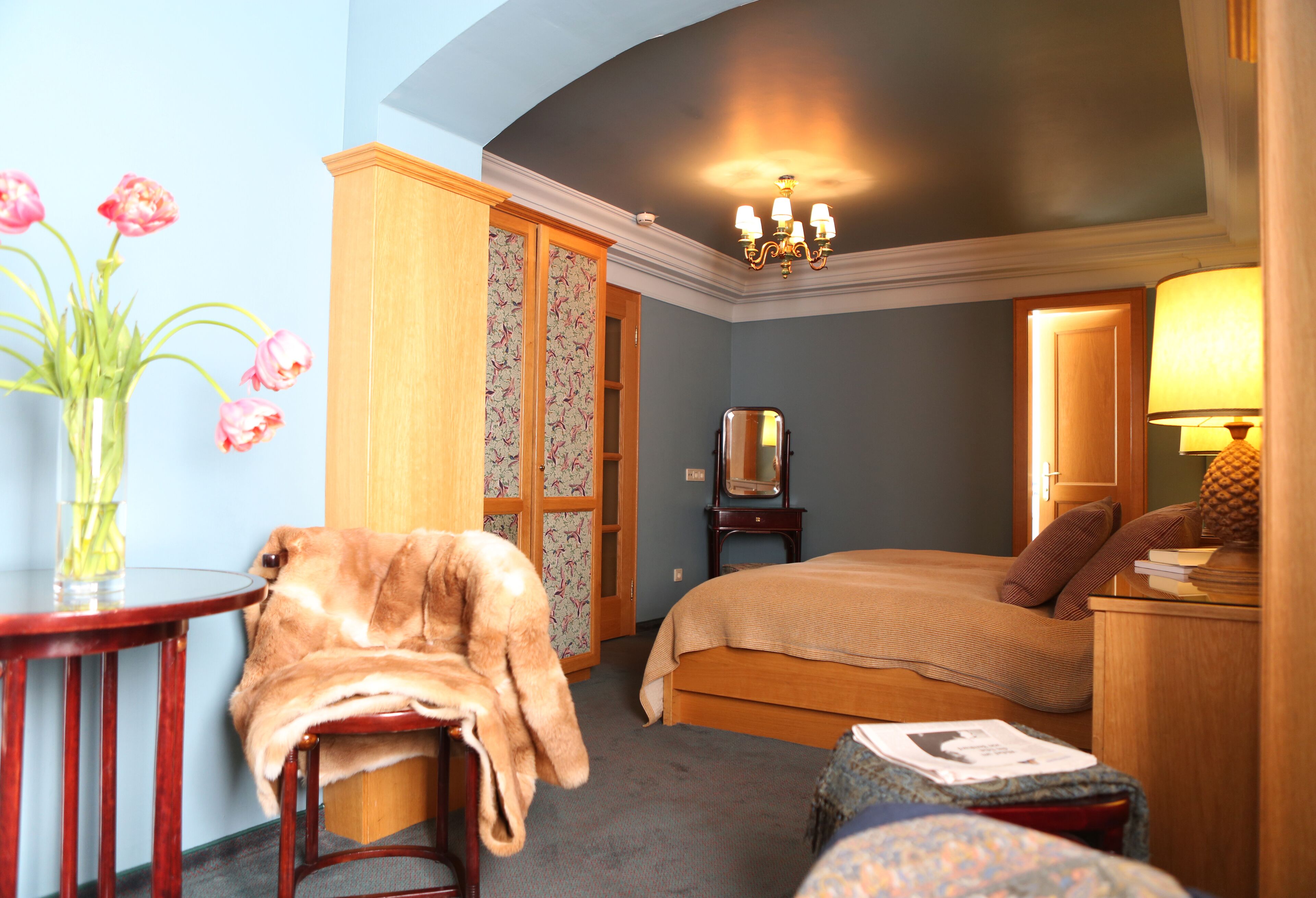 A bedroom with a bed, a bedside table, a lamp, a chair, a small table with a vase of flowers, and a wardrobe.