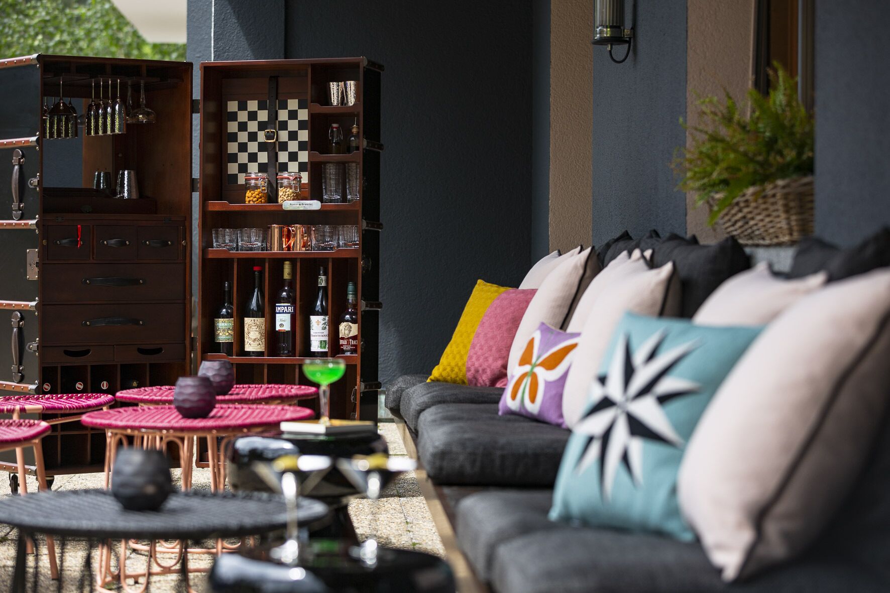 A cozy outdoor seating area with a sofa, cushions, a chessboard, and a bar area with bottles and glasses.