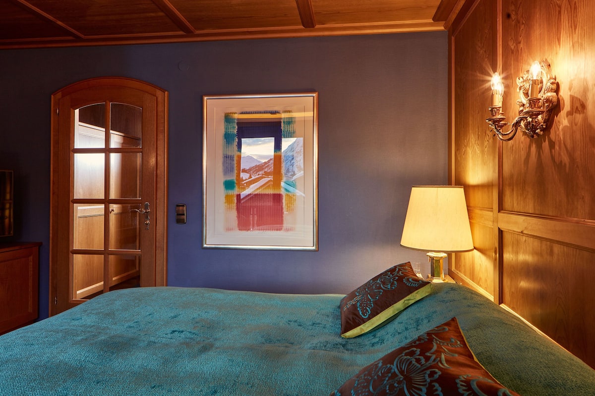 Une chambre à coucher avec un lit, une œuvre d’art encadrée, une porte en bois et un luminaire fixé au mur.