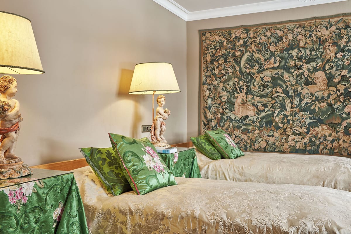 Une chambre d’hôtel avec deux lits, des couvre-lits à motifs floraux verts, une grande tapisserie murale à motifs floraux et une statue sur la table de chevet.