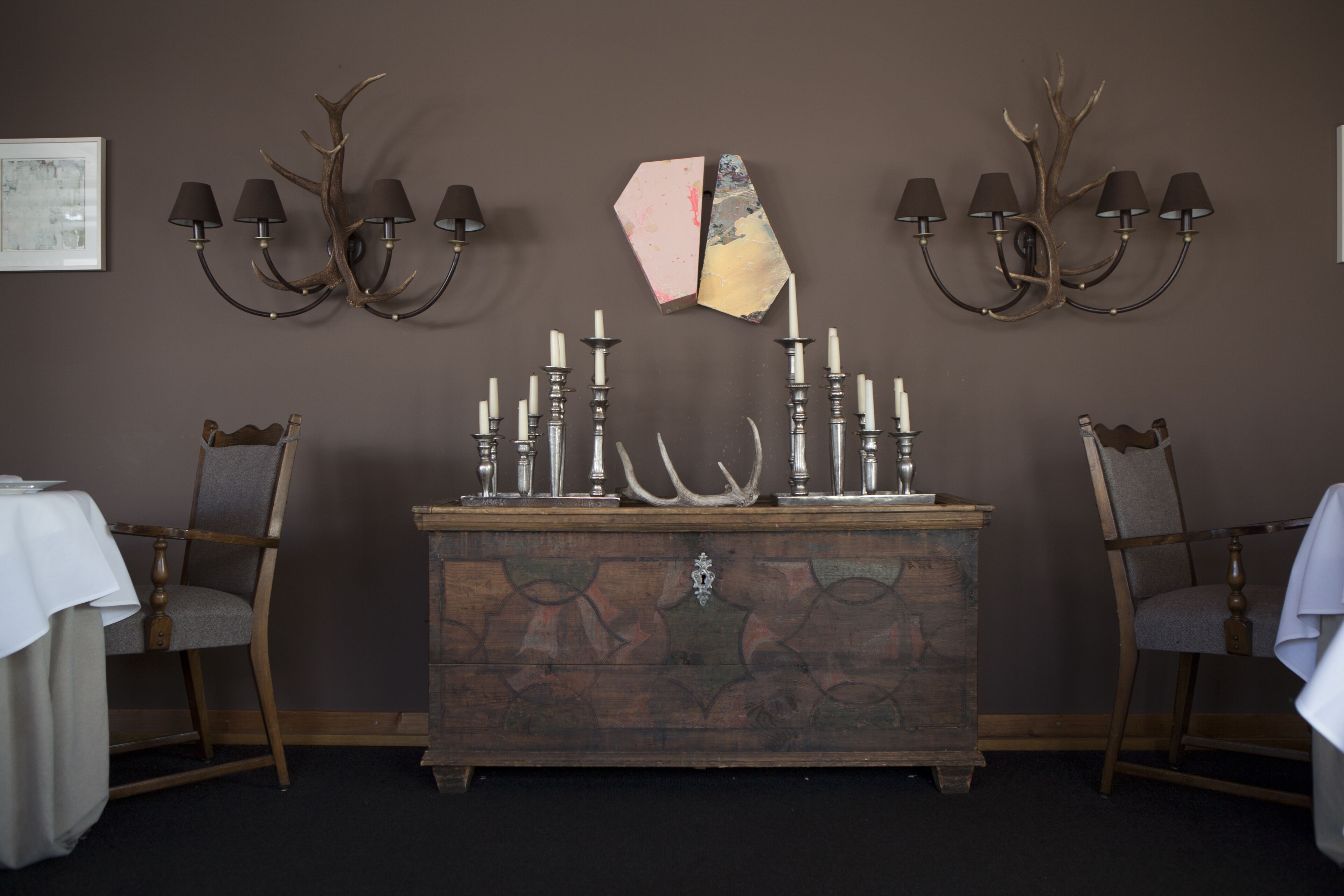 A room with a wooden dresser, two chairs, a table, and a wall-mounted candelabra with candles.