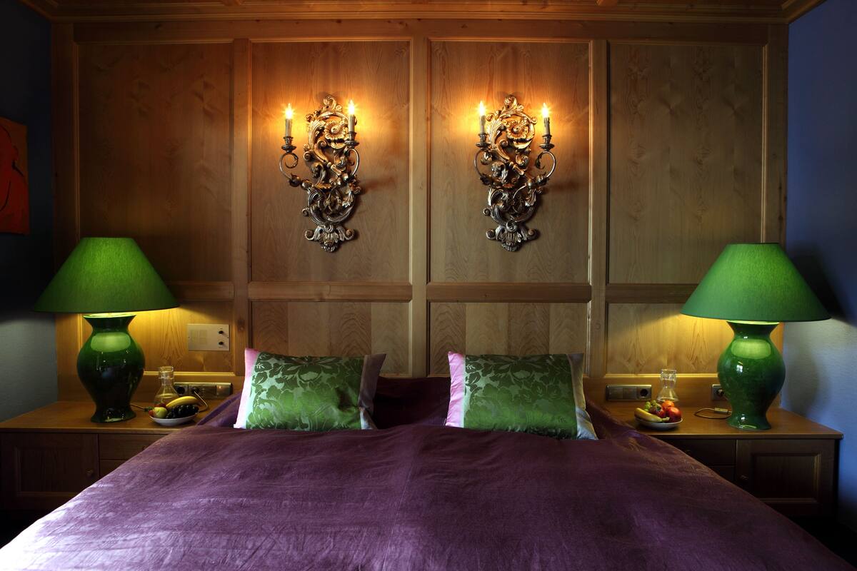 Un lit double avec une couverture violette, deux lampes vertes sur les tables de chevet et un luminaire mural avec des bougies.