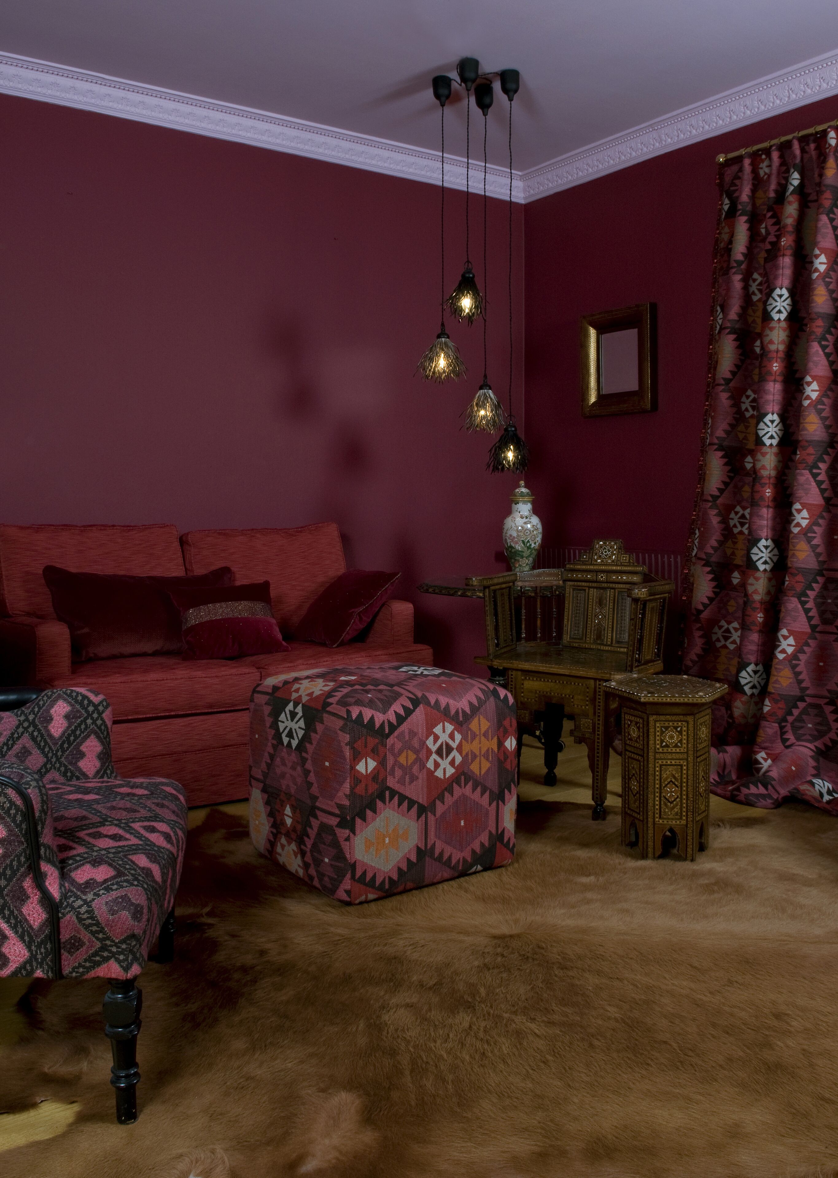 A room with a red sofa, patterned ottoman, a small wooden table, and a patterned curtain.