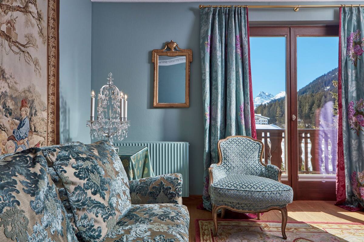 Une pièce avec un canapé à motifs, un fauteuil, un miroir et une vue sur les montagnes par une grande fenêtre.