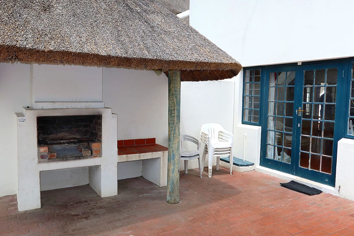 A patio with a thatched roof barbecue, a white wall, a blue door, and a white chair.
