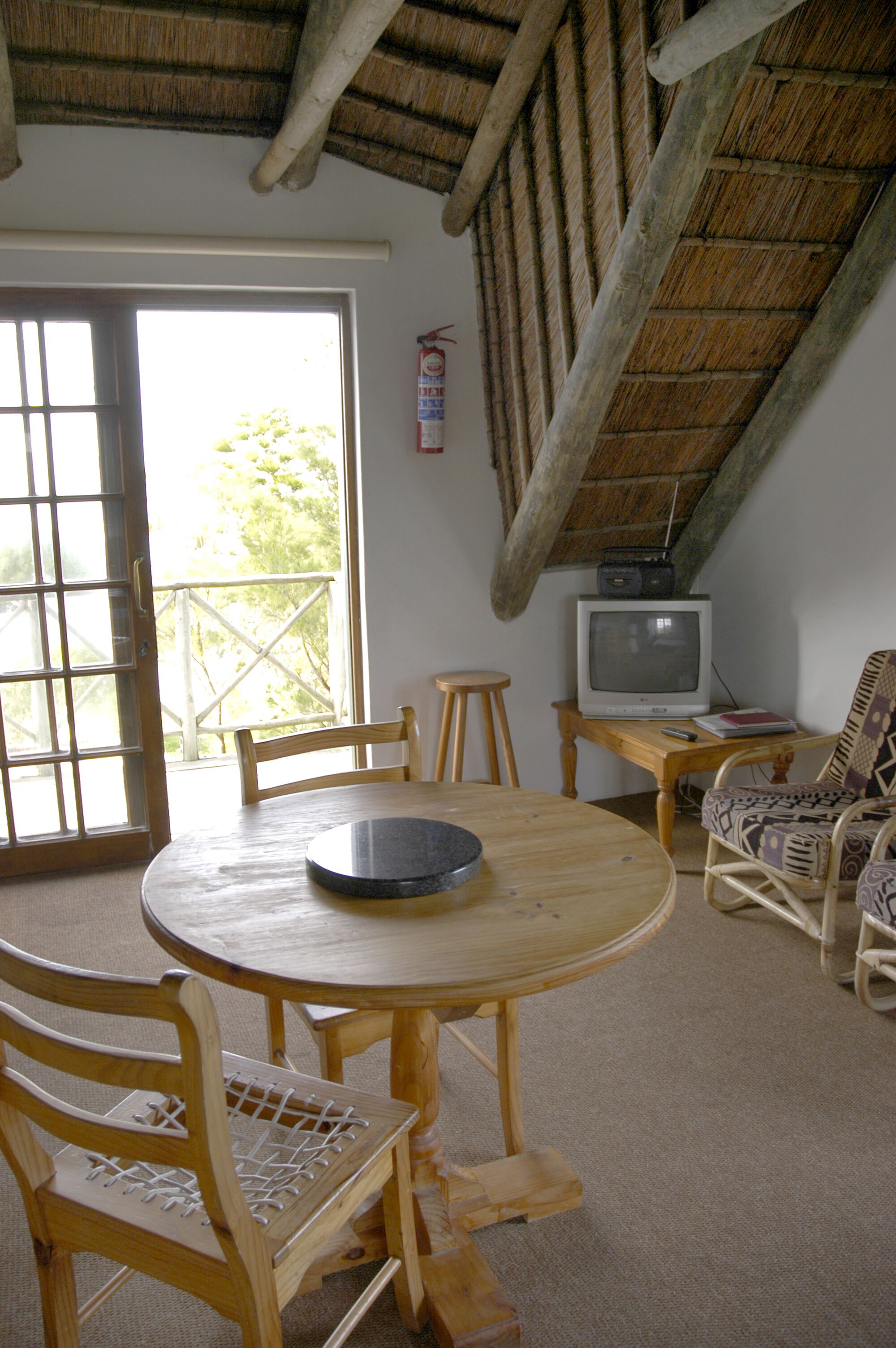 Una habitación acogedora con una mesa redonda de madera, dos sillas de madera, un televisor sobre una mesita y un sillón de mimbre.