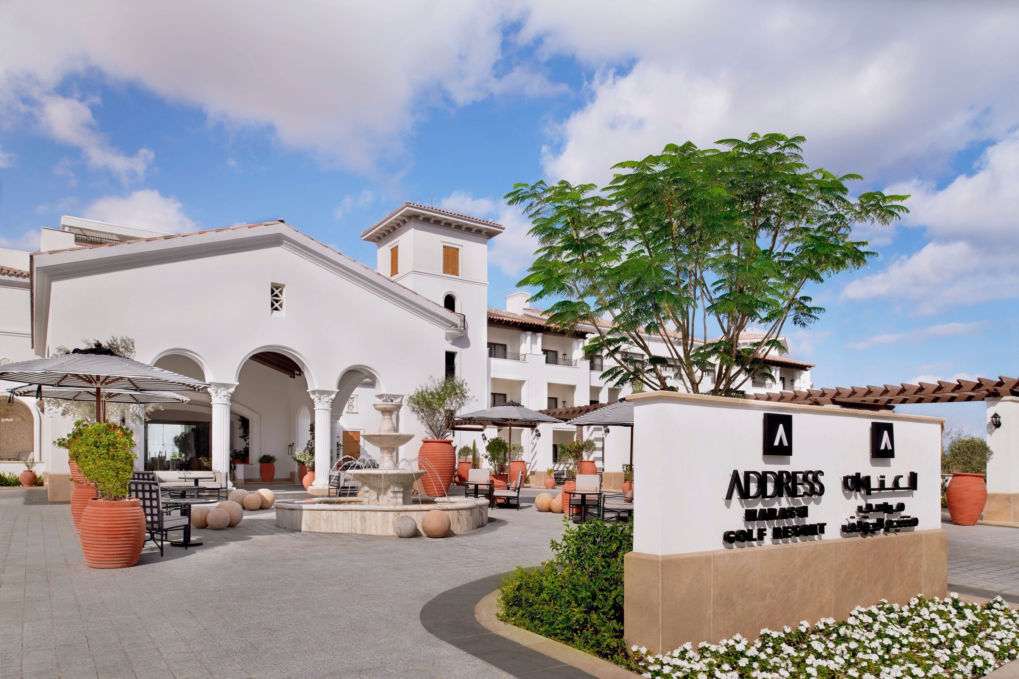 A hotel entrance with a sign, outdoor seating, and a fountain.