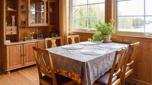 Een eethoek met een tafel voor vier personen, omringd door houten stoelen. Links bevindt zich een houten kast met glazen deuren, en achter de tafel is een raam met uitzicht op bomen.