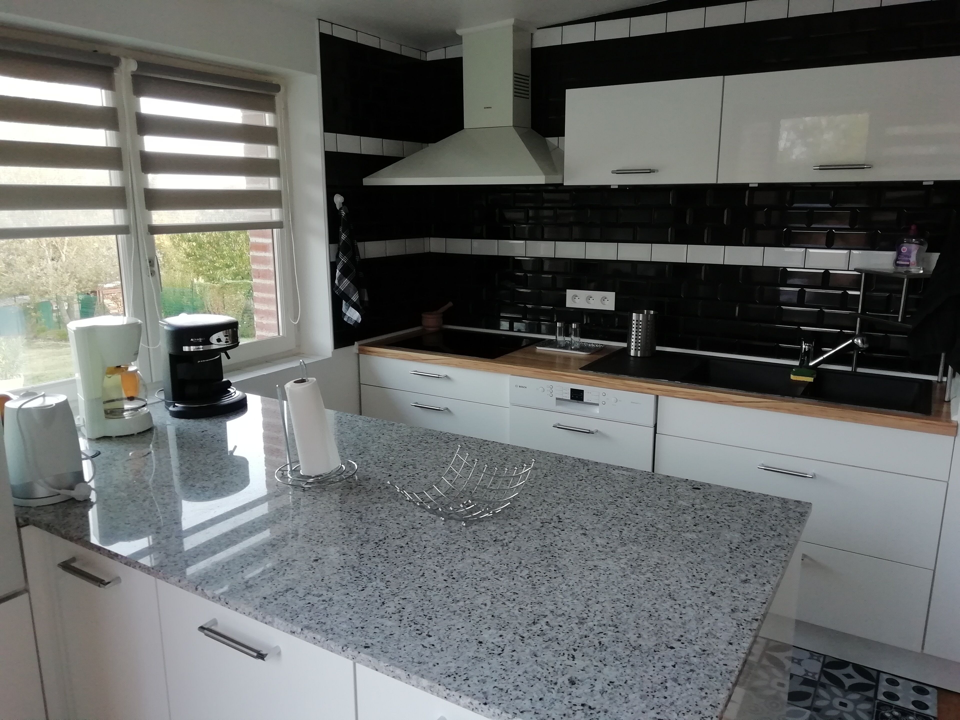 Et moderne køkken med hvide skabe, en sort-hvid backsplash, en granitbordplade og et vindue med persienner.