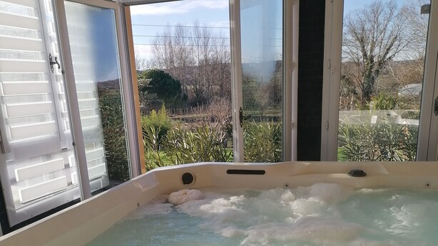 A hot tub with two glasses of champagne and a bottle, a Buddha statue, and a view of trees through open windows. - Gites du Moulin - Le Charmant (Lagardelle-sur-Lèze)