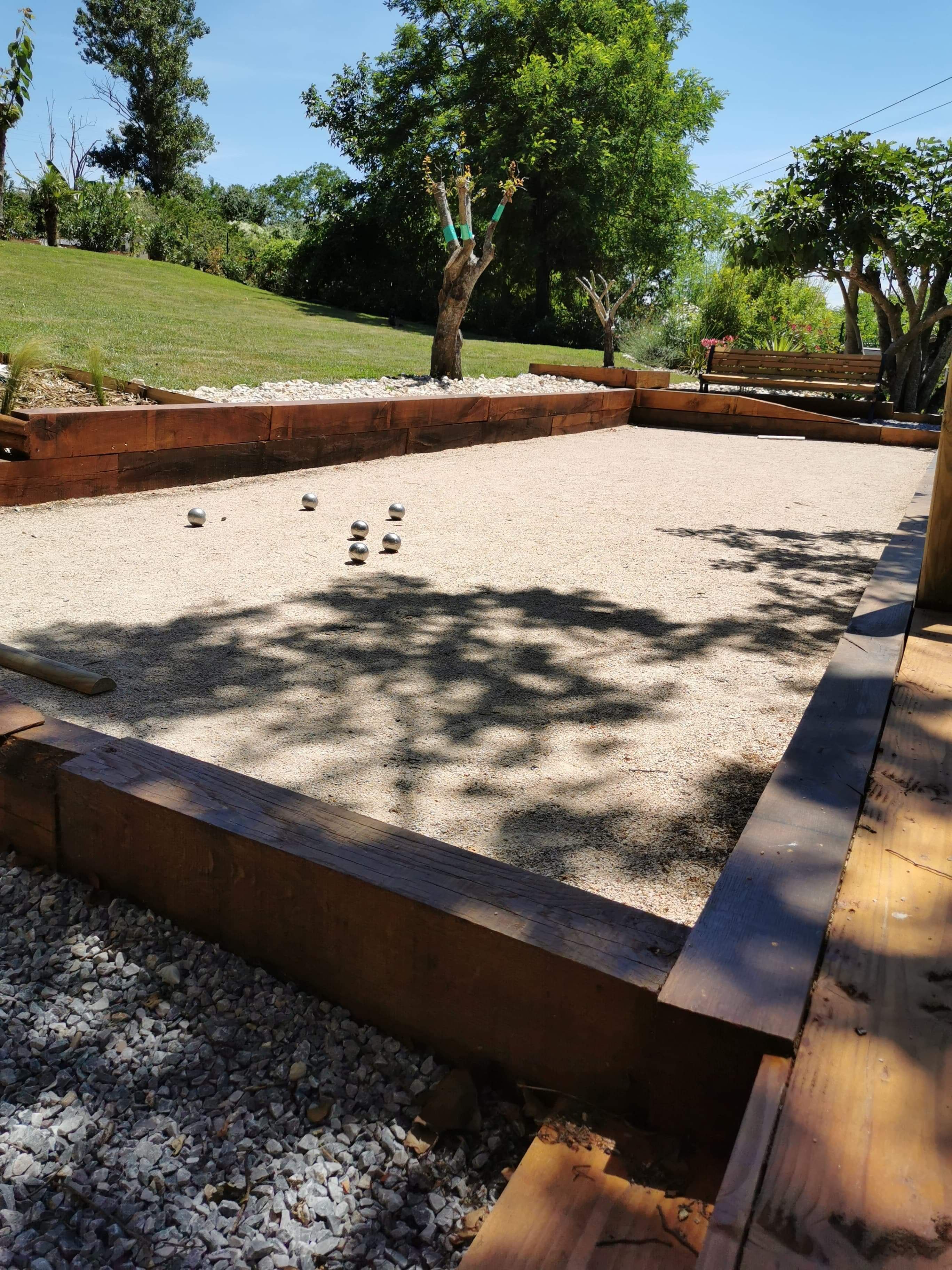 A gravel court with a wooden border, surrounded by a landscaped garden with trees and a bench.