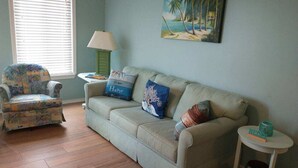 A living room with a beige sofa, a patterned armchair, a small table with a lamp, and a painting of a beach scene. - Wide, Sandy Beaches Await You! (Atlantic Beach)