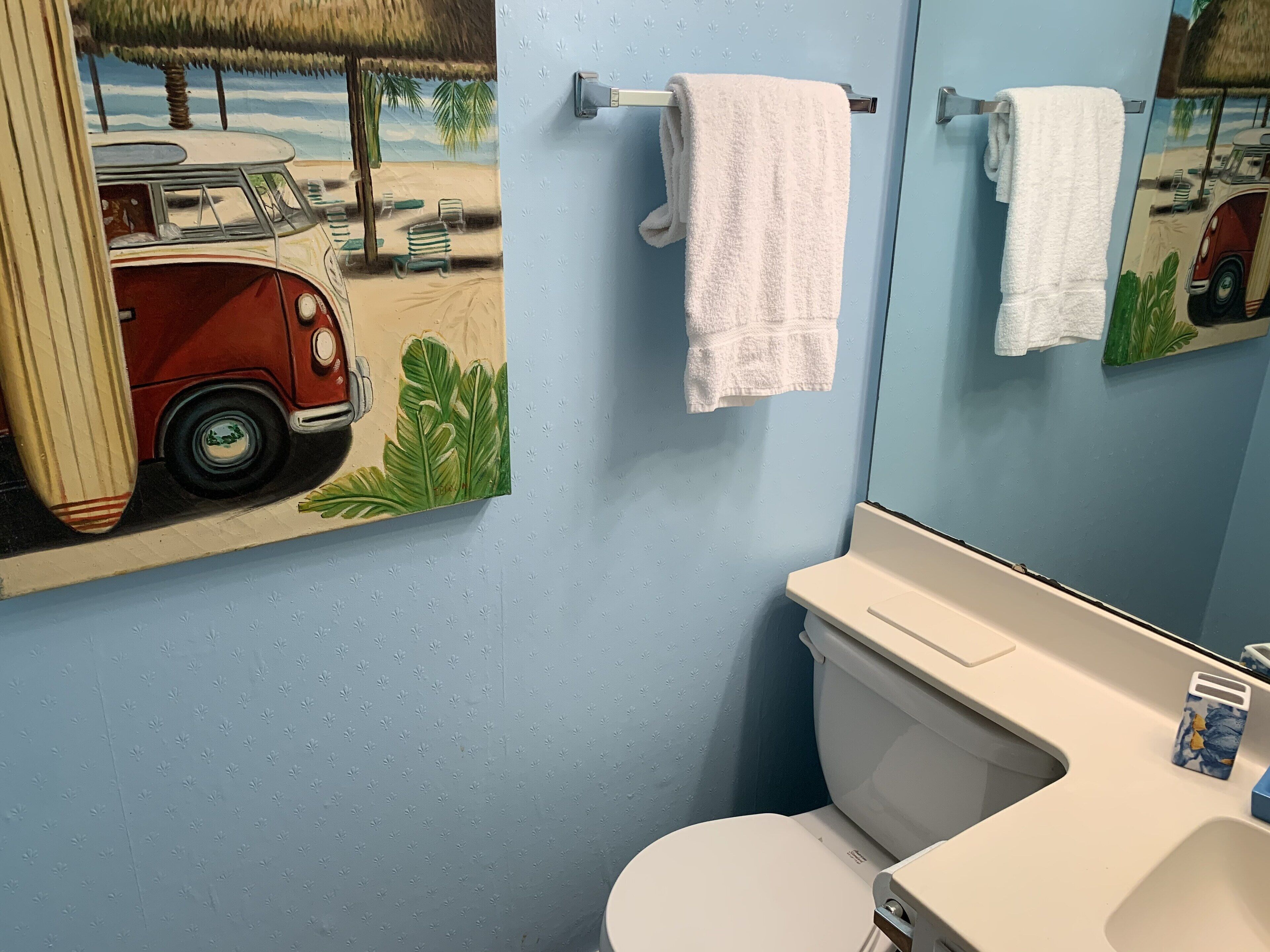 Een badkamer met een toilet, een wastafel en een spiegel. Een handdoek hangt aan de muur en er hangt een schilderij van een auto en palmbomen.