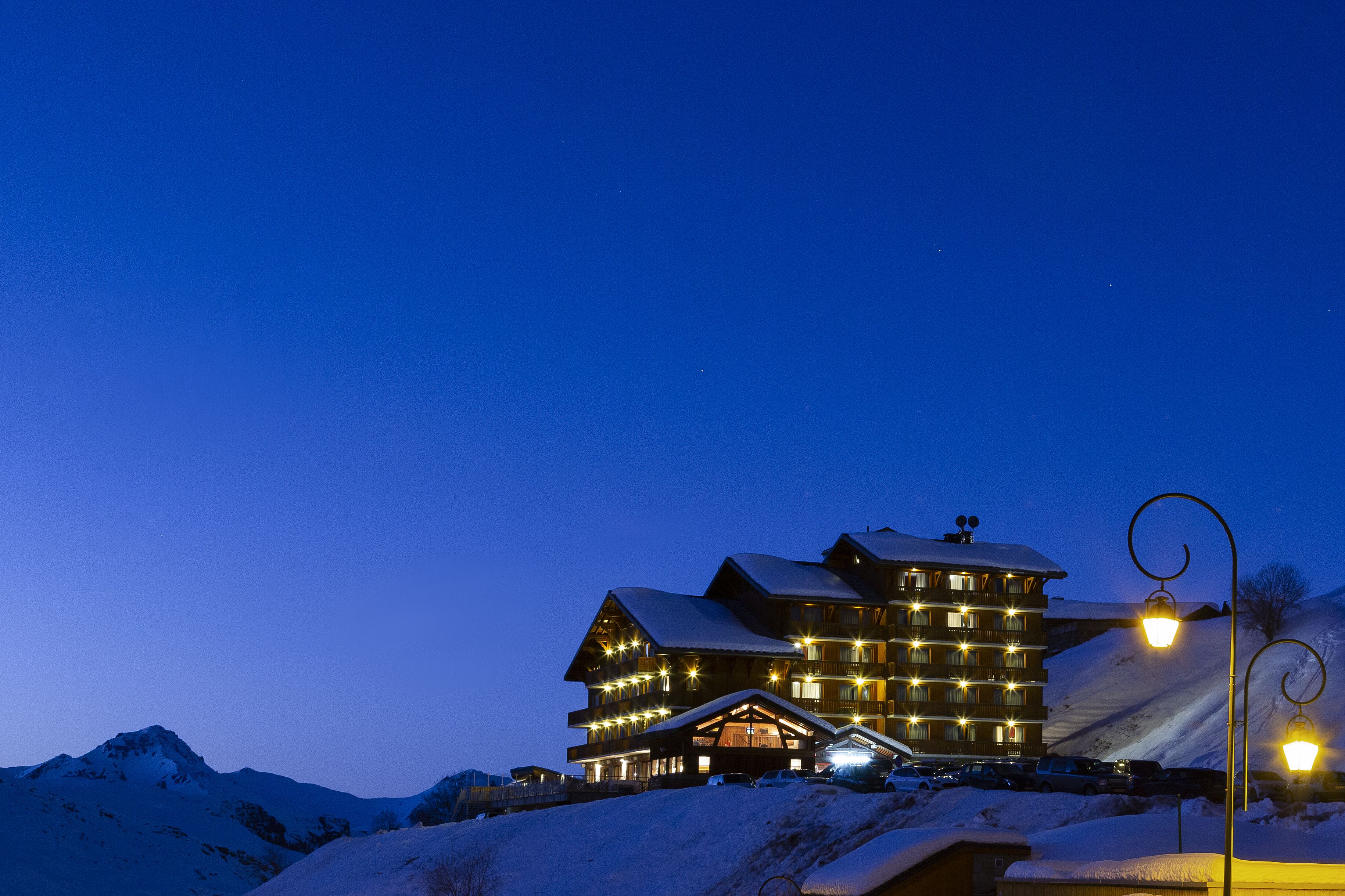 A multi-story building with a snow-covered roof, illuminated windows, and a ski slope in the background.
