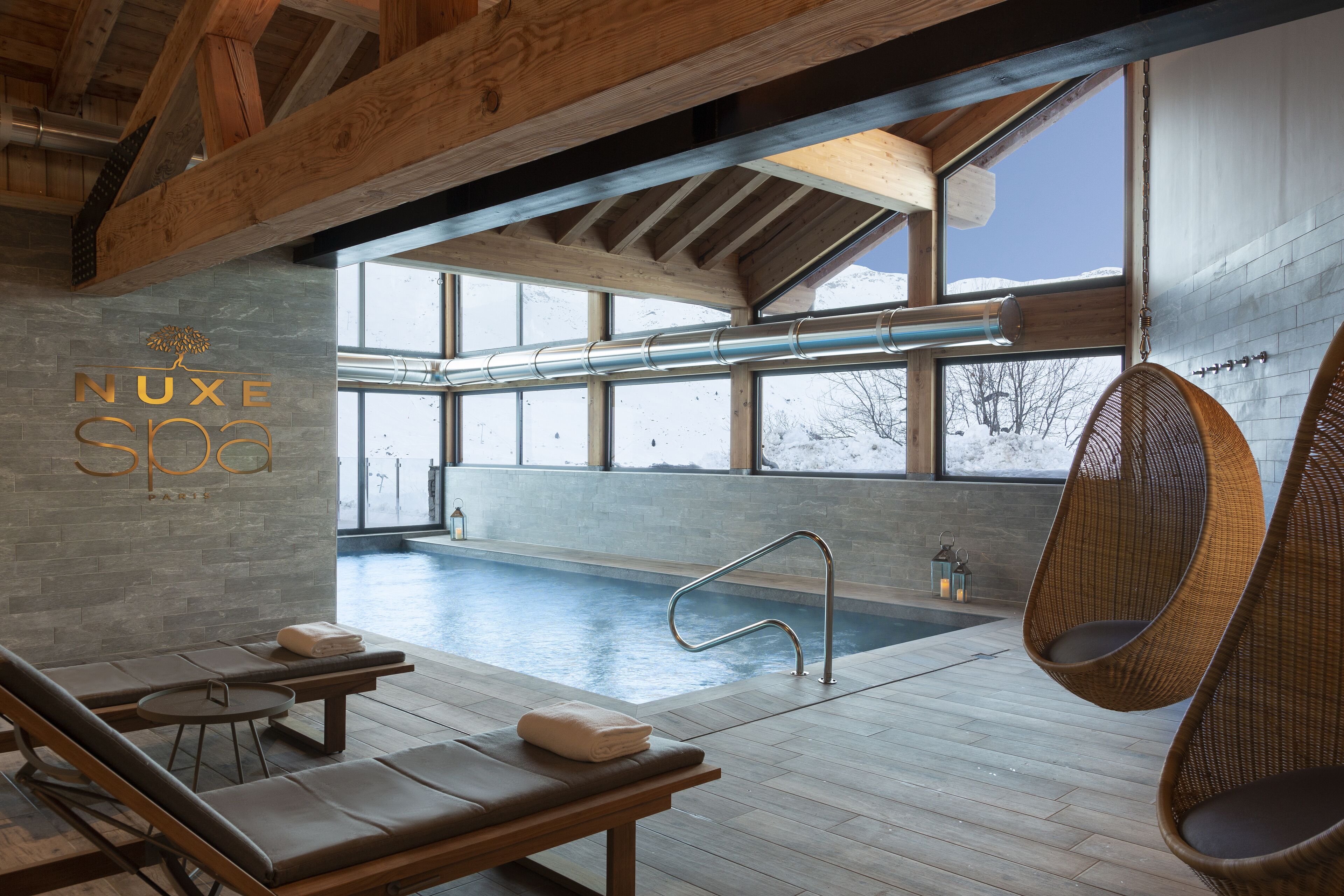 A modern indoor pool area with wooden beams, lounge chairs, and a clear view of the outdoors.