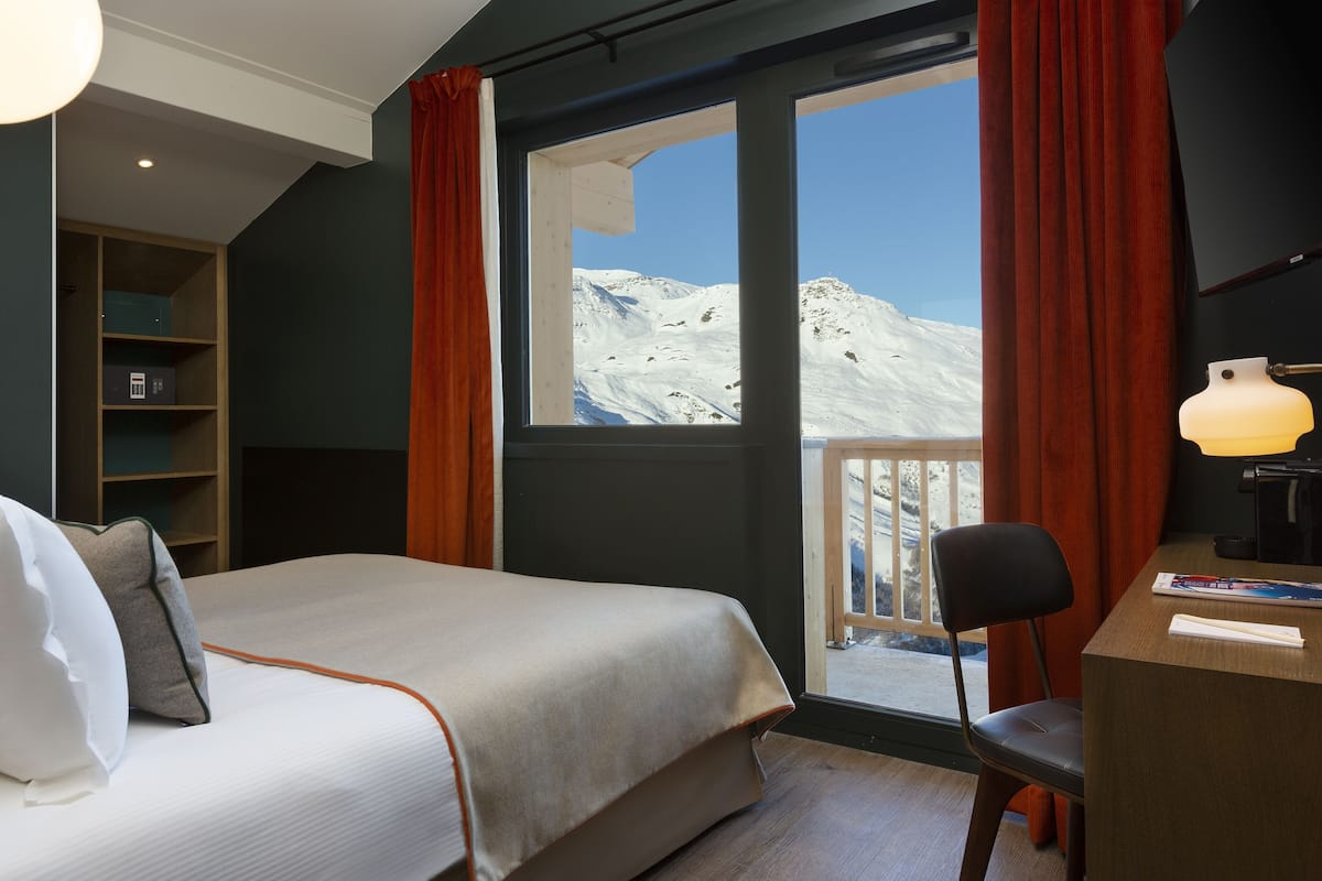 Ein Hotelzimmer mit Bett, Schreibtisch, Stuhl, Fenster mit Blick auf schneebedeckte Berge und einem Bücherregal.