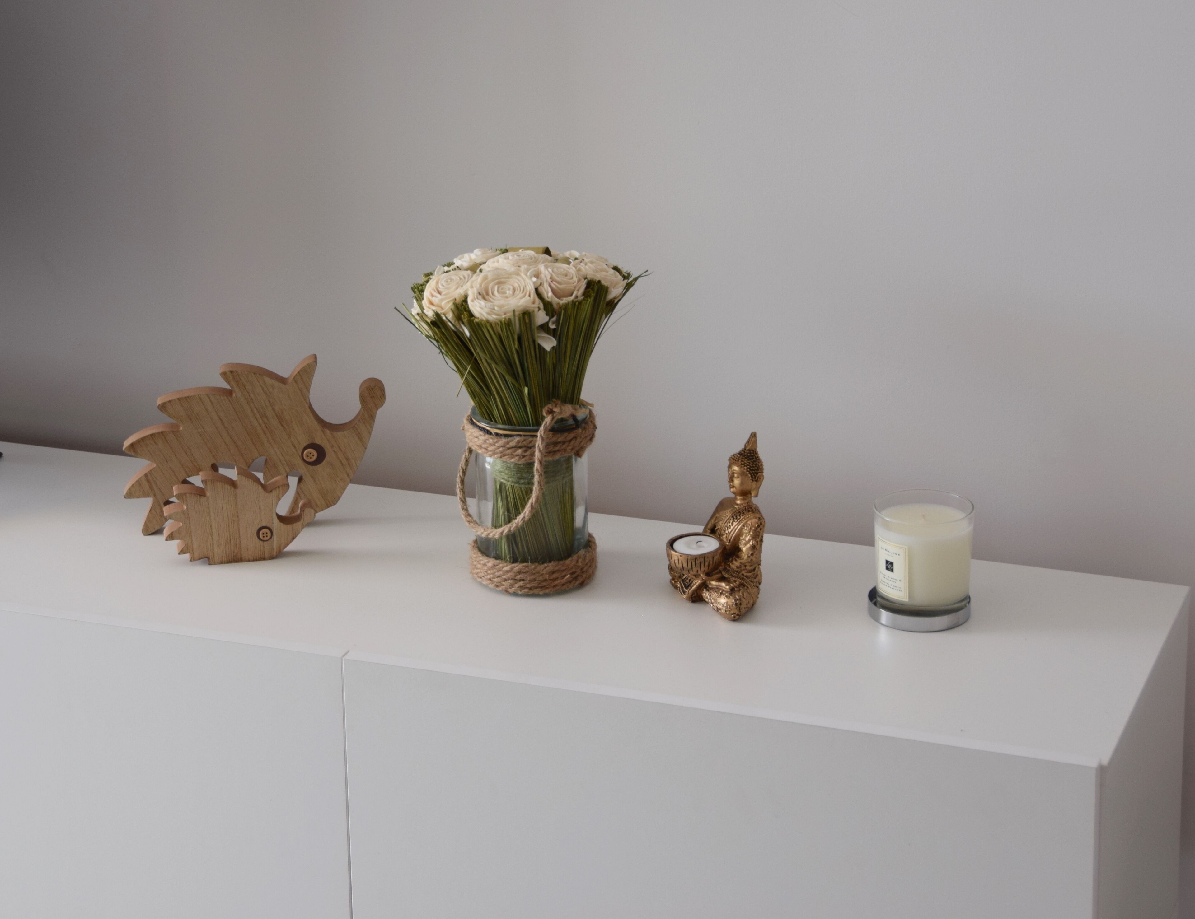 Un mueble blanco con un erizo de madera decorativo, un florero con flores blancas, una pequeña estatua de Buda y una vela.