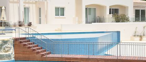 A swimming pool with a wooden deck and metal railing, surrounded by a white building with balconies and windows.