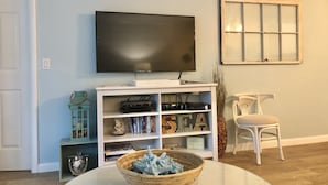 Uma sala de estar com uma TV de tela plana em um móvel, estante, cadeira branca e uma tigela decorativa sobre uma mesa.
