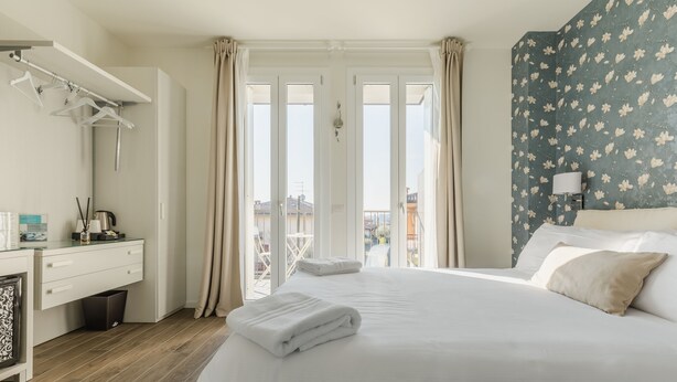 A hotel room with a bed, a desk with a lamp, a trash bin, and a view of the city through large windows. - Casa Baci (Lazise)