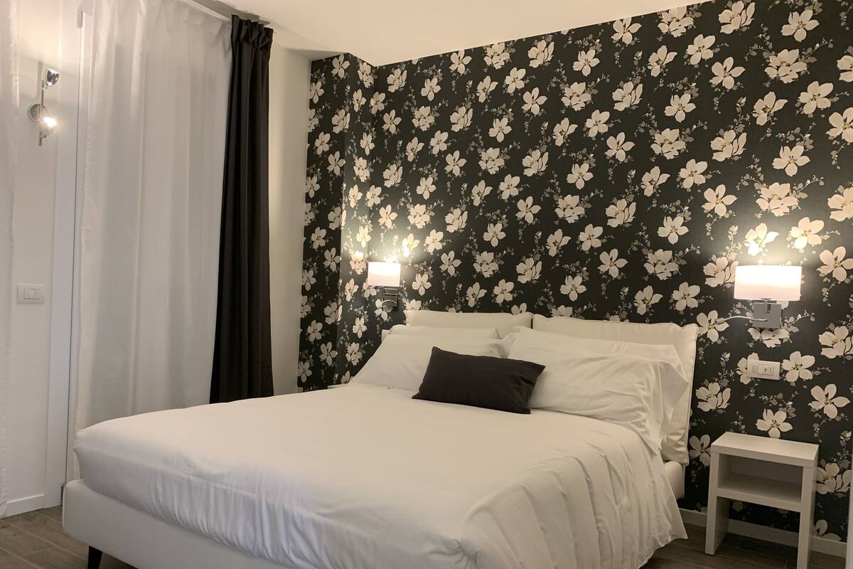 Una habitación de hotel con una cama, dos mesitas de noche, una lámpara de pared y un ventanal con cortinas.