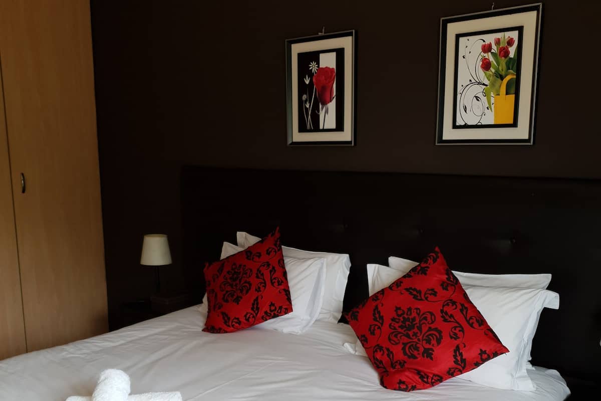 A neatly made double bed with white linens and red patterned pillows, a wooden wardrobe, and two framed pictures on the wall.
