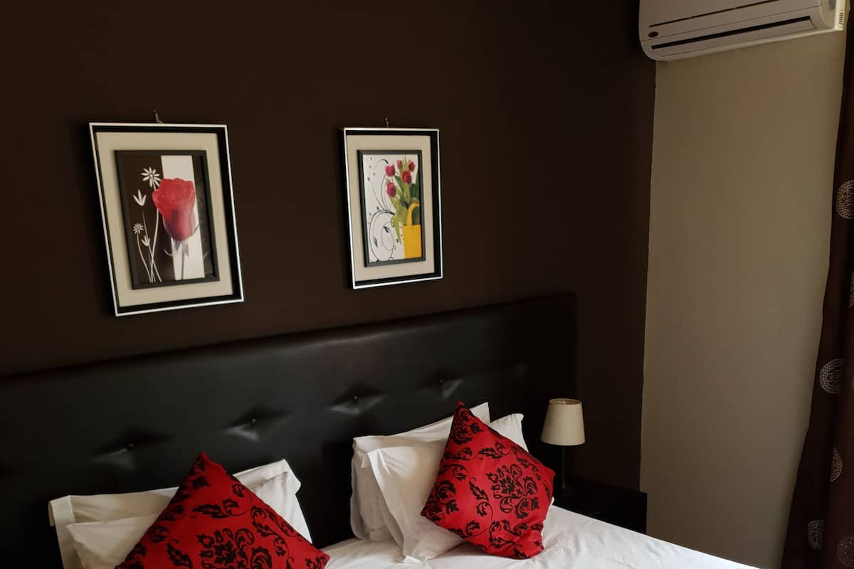A hotel room with a large bed, two framed floral artworks on the wall, a dark headboard, and an air conditioning unit.
