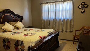Una camera da letto con un letto grande, una testiera in legno, una finestra con tende leggere, una sedia di legno e una croce appesa al muro.