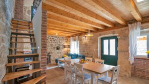 A rustic interior with exposed wooden beams, a stone wall, a dining area with white chairs, and a staircase with wooden steps.
