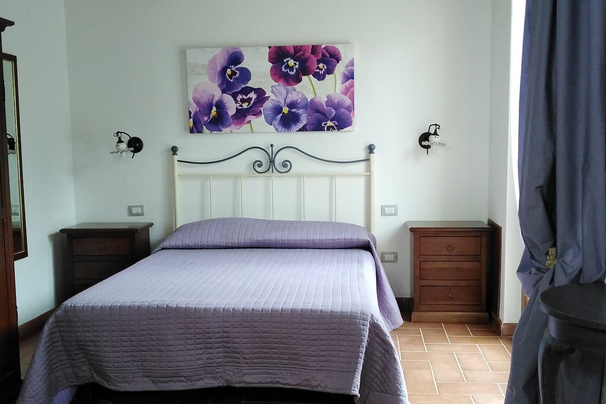 Een slaapkamer met een bed, twee nachtkastjes, een wandlamp, een decoratief bloemen schilderij en een betegelde vloer.