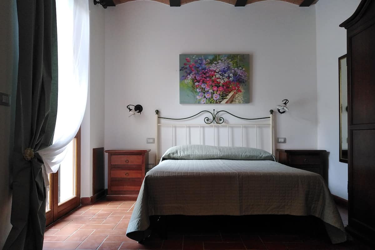 Een slaapkamer met een bed, nachtkastje, dressoir, schilderij en een raam met gordijnen.