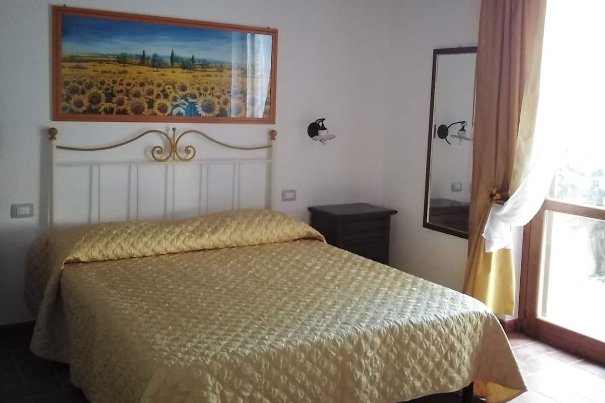 Een slaapkamer met een bed, een schilderij aan de muur, een nachtkastje en een raam met gordijnen.