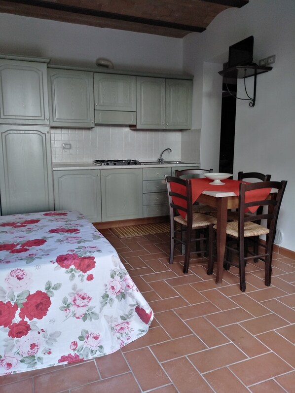 A compact kitchen with a gas stove, sink, and light green cabinets. There is a dining table with four chairs and a red tablecloth. The floor is tiled, and the room has a simple, functional design.