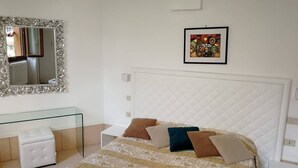 Un dormitorio con una cama blanca, una mesita de noche de cristal, una silla blanca, un espejo con marco decorado y un cuadro en la pared.