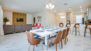 Uma área de jantar moderna com uma mesa branca posta para seis pessoas, rodeada por cadeiras de madeira com almofadas azuis.