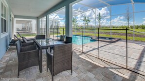 Alpendre coberto com mesa de vidro e cadeiras de vime, com vista para uma piscina e área ajardinada.