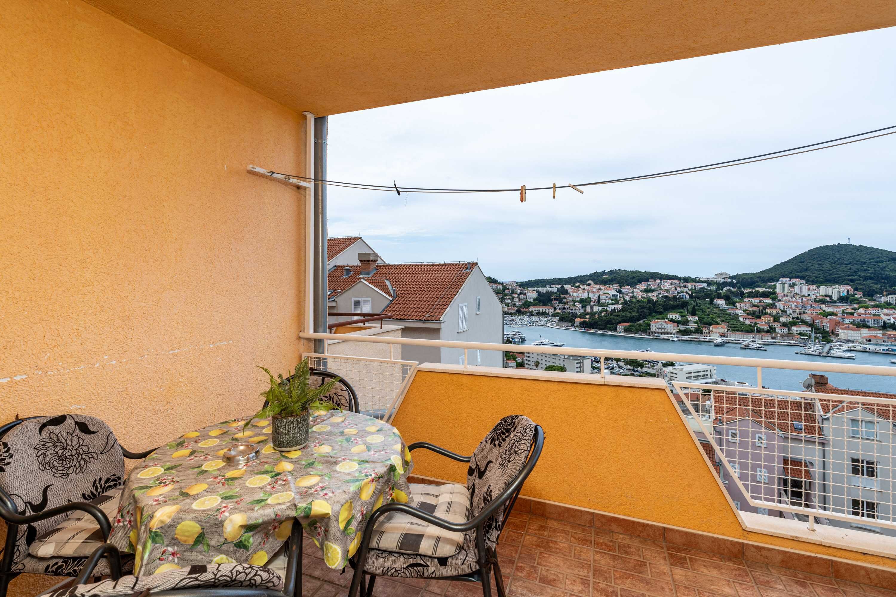 A balcony with a table and chairs, a potted plant, and a view of a coastal town and marina.