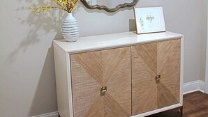 Una consola blanca con un mueble de diseño geométrico, un jarrón con flores amarillas y un certificado enmarcado encima.