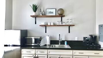 Una cocina equipada con microondas, cafetera y lavamanos. Hay dos estantes sobre la encimera con diversos objetos.