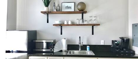 Una cocina equipada con microondas, cafetera y lavamanos. Hay dos estantes sobre la encimera con diversos objetos.