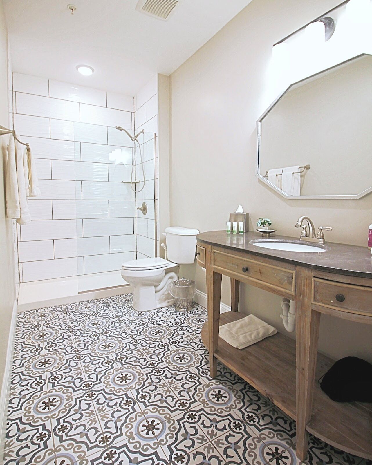 A bathroom with white tiled walls, a patterned floor, a wooden vanity, and a shower.