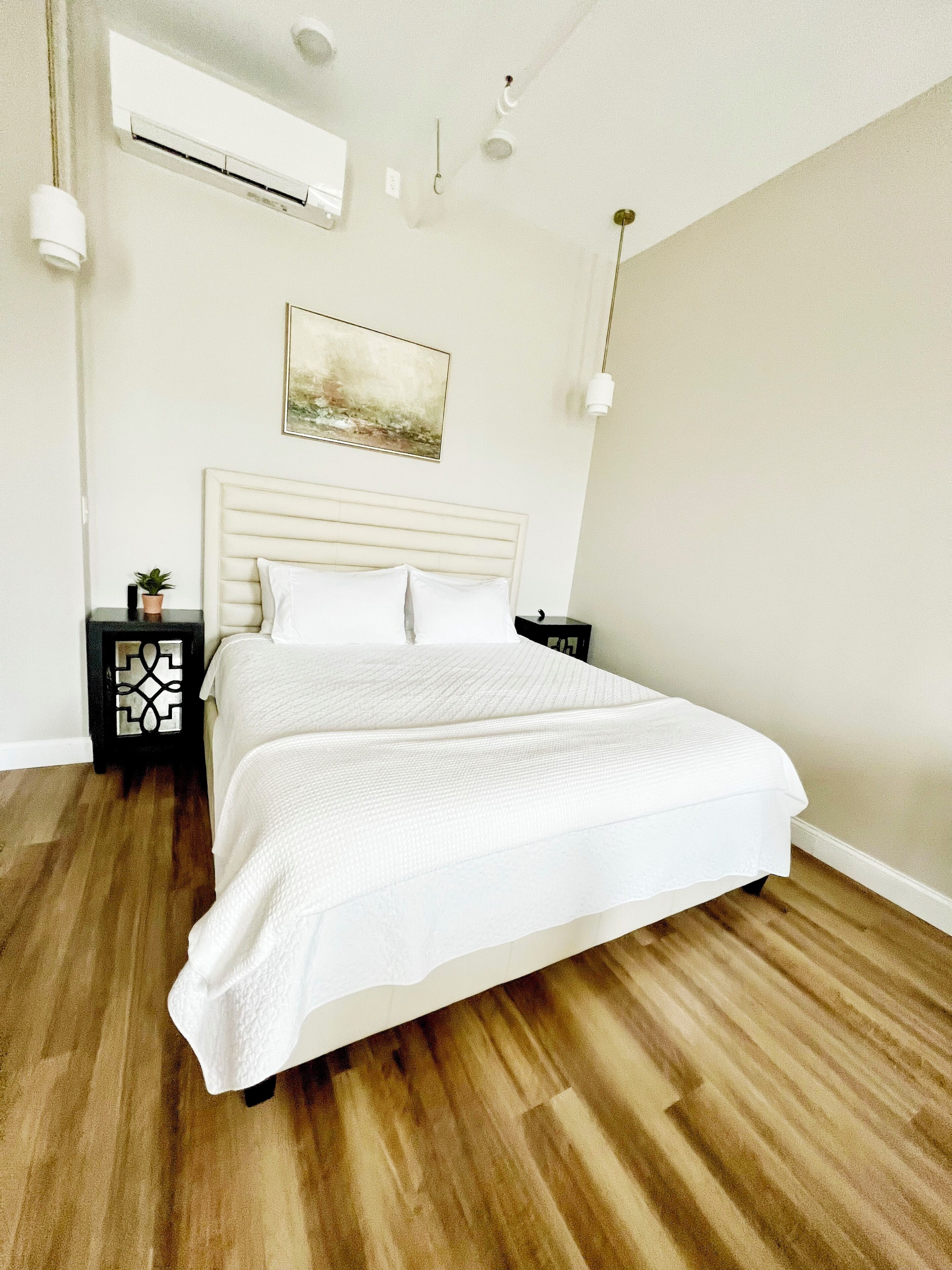 A bedroom with a large bed, a headboard, a bedside table with a plant, and a wall-mounted air conditioner.