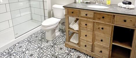 Baño con ducha, inodoro, tocador y baldosas de suelo estampadas.
