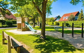 Een speelplaats met een glijbaan en klimgebeuren, omringd door bomen en een bank.