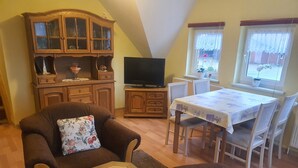 Een gezellige woonkamer met een bruine bank, een kleine tafel met een afstandsbediening en een kaars, een televisie op een houten kast, en een eethoek met een tafel en stoelen.