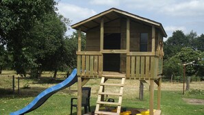 Een houten speelhuisje met een glijbaan en een ladder die leidt naar een platform met speelvoertuigen.