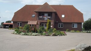 Een gebouw met twee verdiepingen, een dak van rode tegels, een overdekte parkeerplaats en een aangelegde tuin met verschillende planten en bloemen.