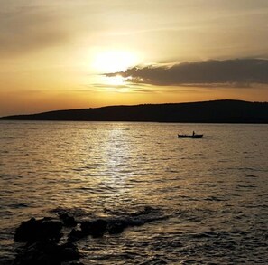 Un coucher de soleil sur une étendue d’eau calme, avec la silhouette d’un bateau et une terre lointaine à l’horizon.
