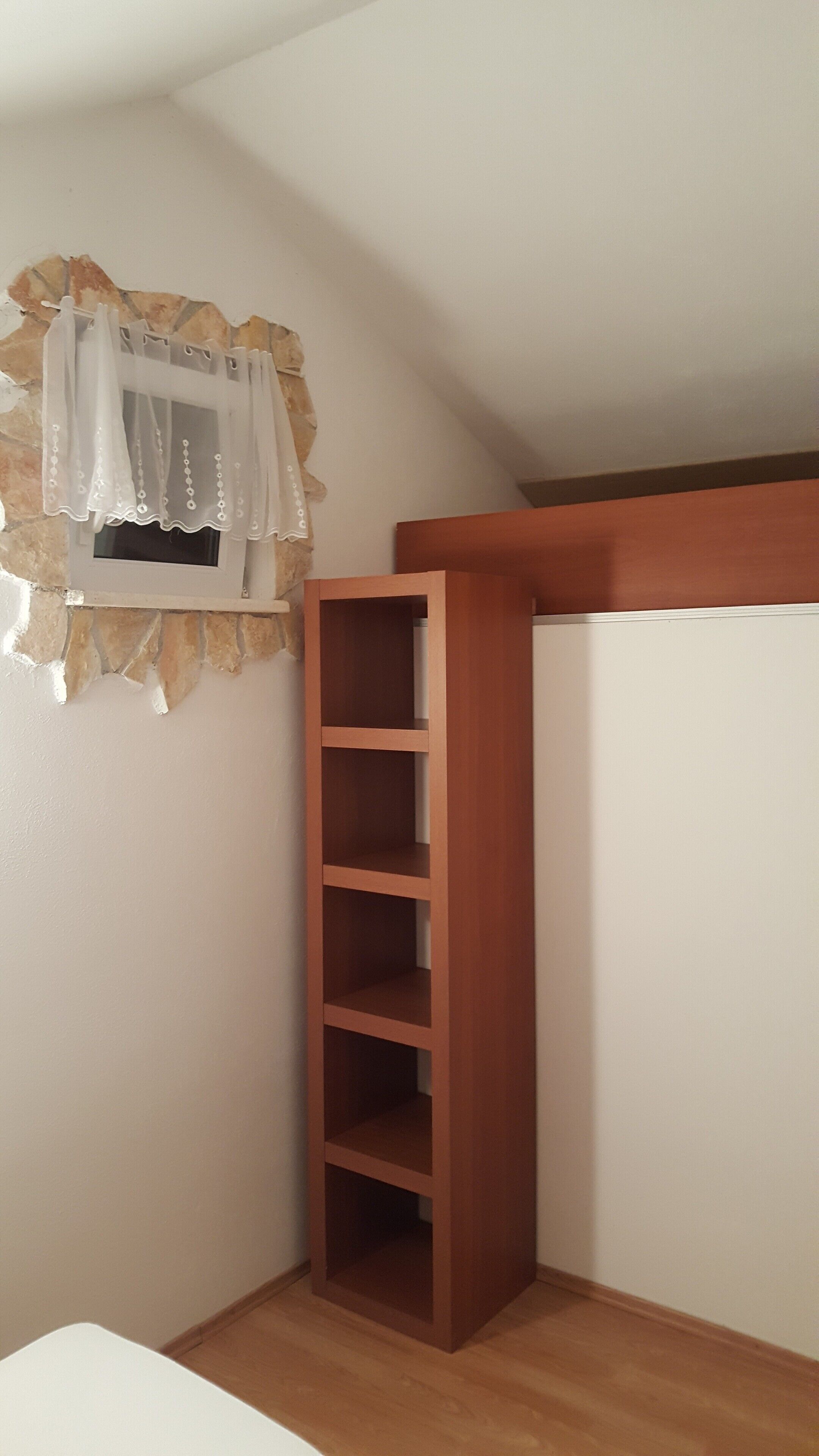 Una camera d'angolo con un'unità di scaffalatura in legno, una piccola finestra con tenda e una parete bianca.